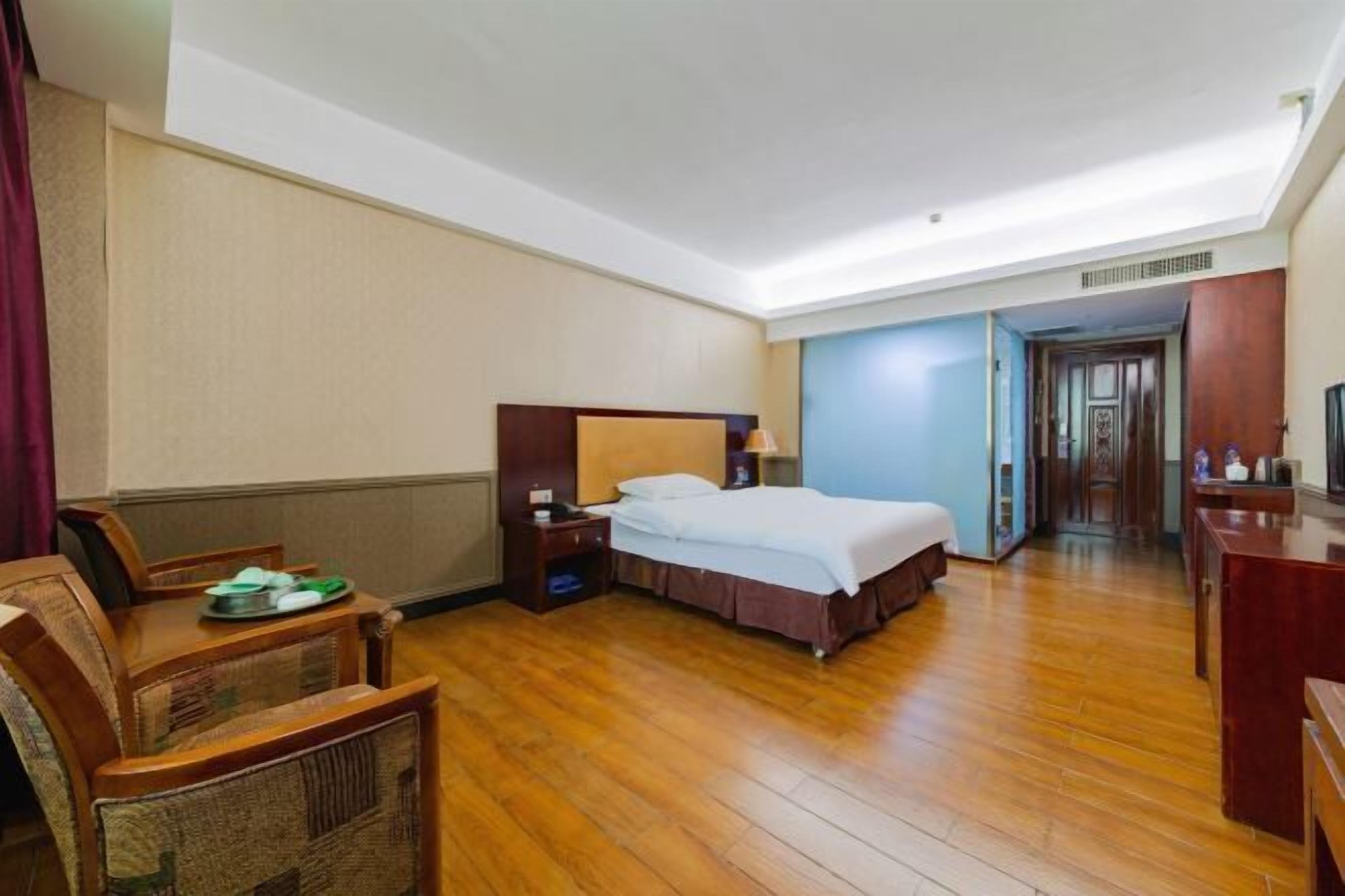 A hotel room with a wooden floor, a bed with white linens, a nightstand, a chair, and a desk with a lamp.