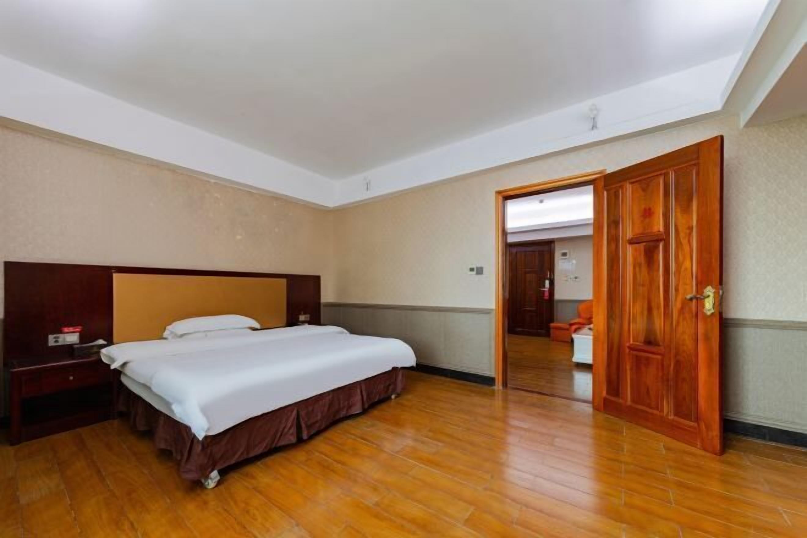 A hotel room with a wooden floor, a bed with a white bedspread, a wooden headboard, and a wooden door.