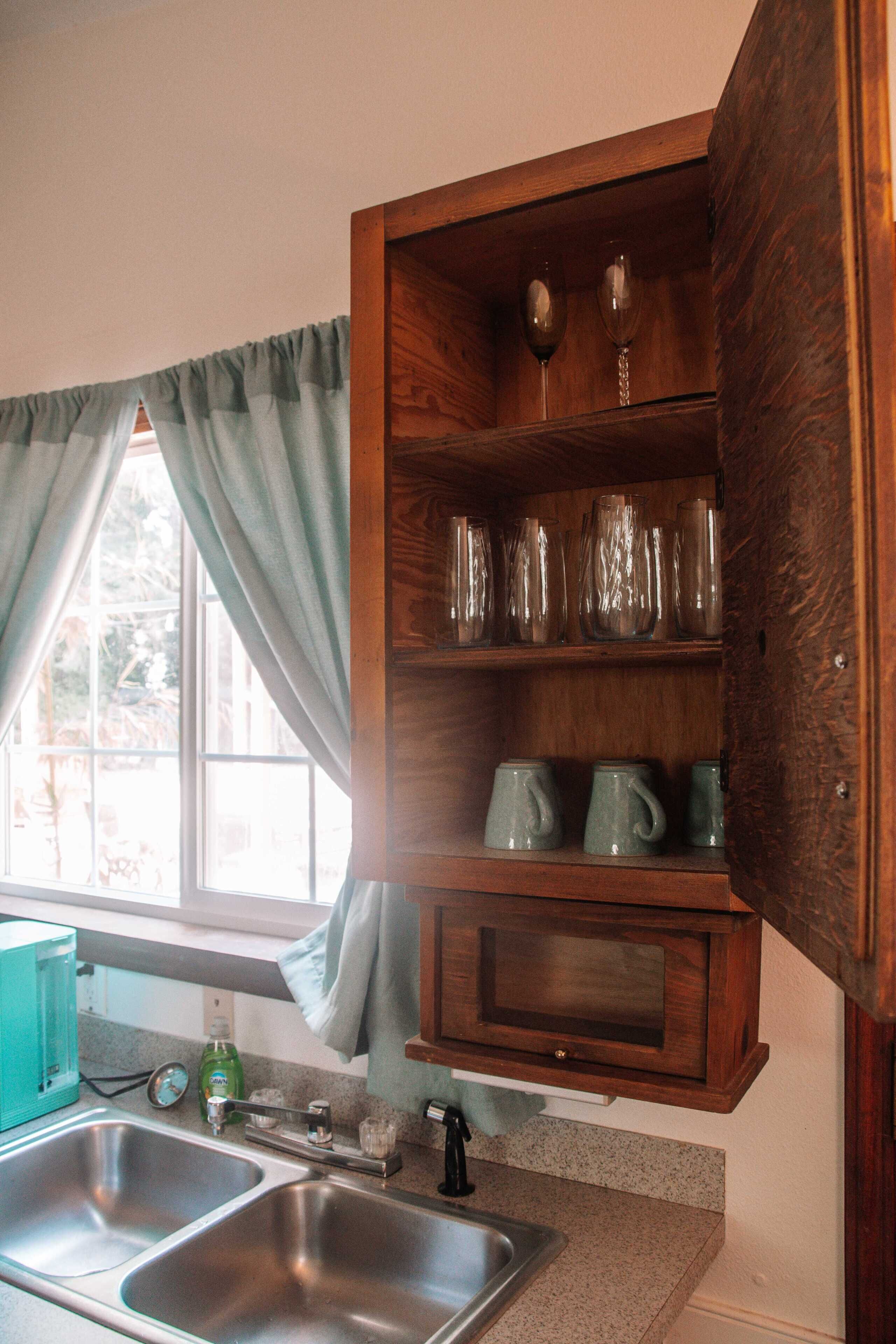 Una cocina con dos fregaderos, una ventana con cortinas y un armario de madera con cristalería.