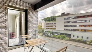 A balcony with a wooden table and chairs, a glass door leading to an interior space, and a view of a modern building across the street.
