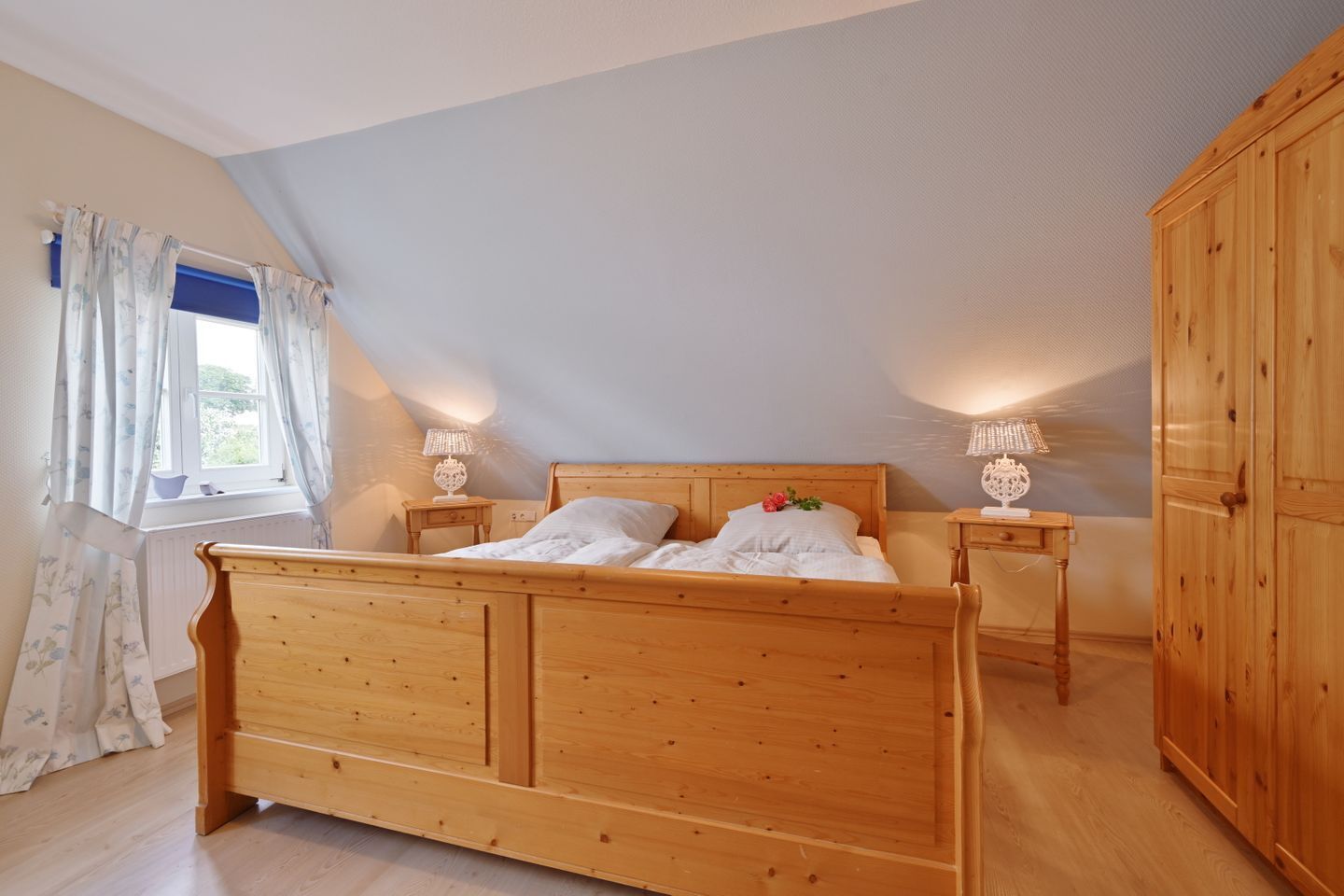 A bedroom with a wooden bed, two bedside tables with lamps, a window with curtains, and a wardrobe.