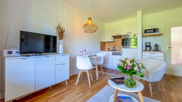 Un apartamento moderno con sala de estar, comedor y cocina. Cuenta con un televisor de pantalla plana, muebles blancos, una mesa de comedor y una cocina equipada con microondas y horno.