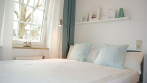 Un dormitorio con cama, ventana, radiador, lámpara y una estantería con fotografías.