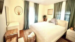 A bedroom with a large bed, a bedside table, a lamp, a fan, a potted plant, and a wicker basket.