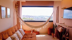 A cozy living room with a hammock, a couch with cushions, a TV, and a view of a cityscape through a large window. - AxéHome | Charming Accommodation | 2 Bedrooms / 2 Bathrooms (Salvador)