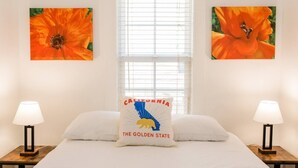 A bedroom with a bed, two nightstands, two lamps, a window with blinds, and two paintings of orange flowers. - 2 BD Quiet Neighborhood with AC, Desk, Fast Wifi (Torrance)