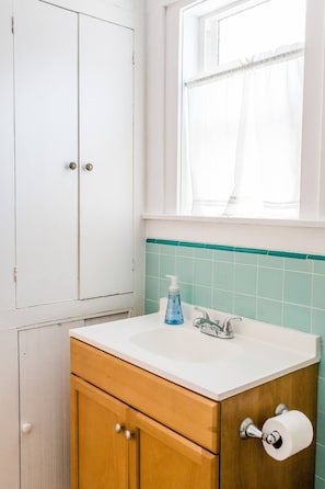 Un baño con lavamanos blanco, armarios de madera, una ventana con cortinas traslúcidas y un salpicadero con azulejos verdes.