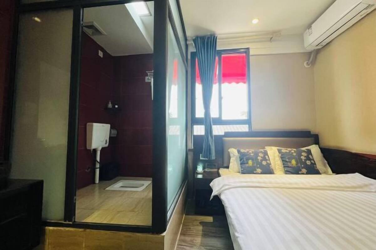 A hotel room with a bed, a nightstand, a window with curtains, a wall-mounted air conditioner, and a wooden floor.
