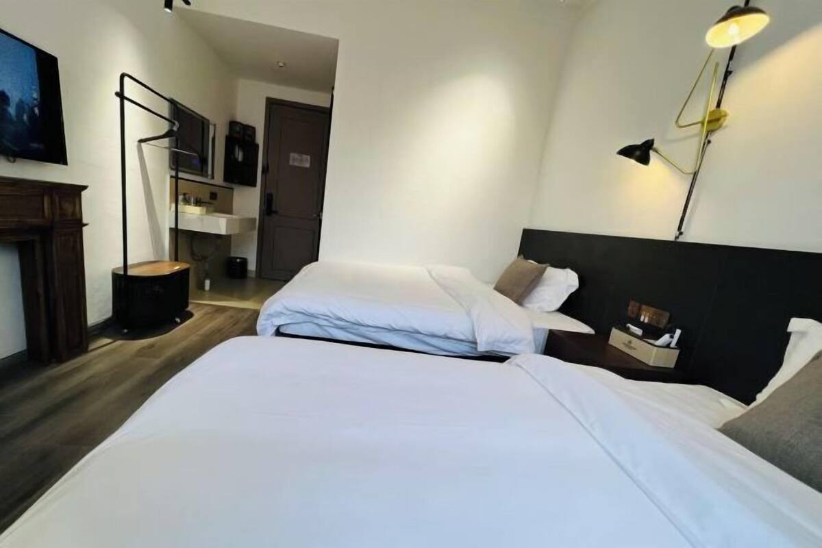 A modern hotel room with a large bed, bedside tables, a desk, and a wall-mounted lamp.