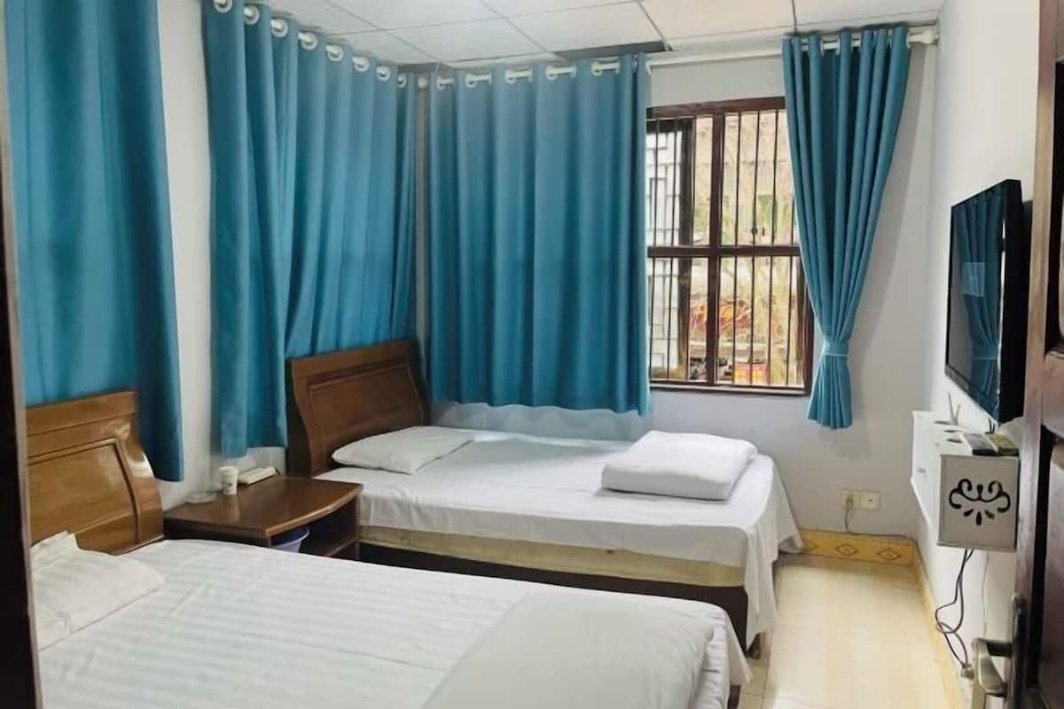 A hotel room with two beds, a wooden headboard, a small bedside table, a TV, and a window with blue curtains.