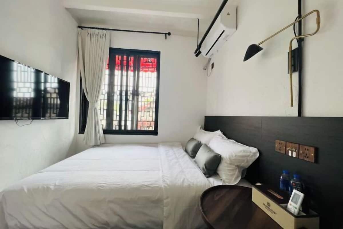 A hotel room with a bed, a TV mounted on the wall, an air conditioning unit, a desk with a chair, and a window with curtains.