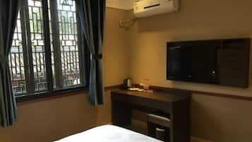 A hotel room with a bed, a desk with a chair, a television mounted on the wall, and an air conditioning unit.