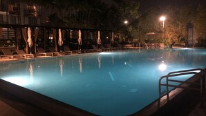 A nighttime swimming pool with lounge chairs and umbrellas, surrounded by palm trees and buildings.