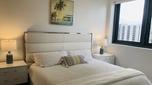 A neatly made bed with white linens and multiple pillows, flanked by two bedside tables with lamps. A painting of a beach scene hangs above the bed.