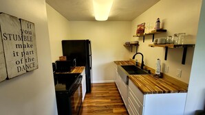Une cuisine compacte avec des plans de travail en bois, un réfrigérateur noir et une étagère murale où sont rangées la vaisselle.