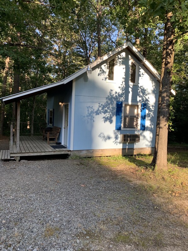 Une petite cabane à un seul étage, dotée d’un porche et d’une cheminée, entourée d’arbres.