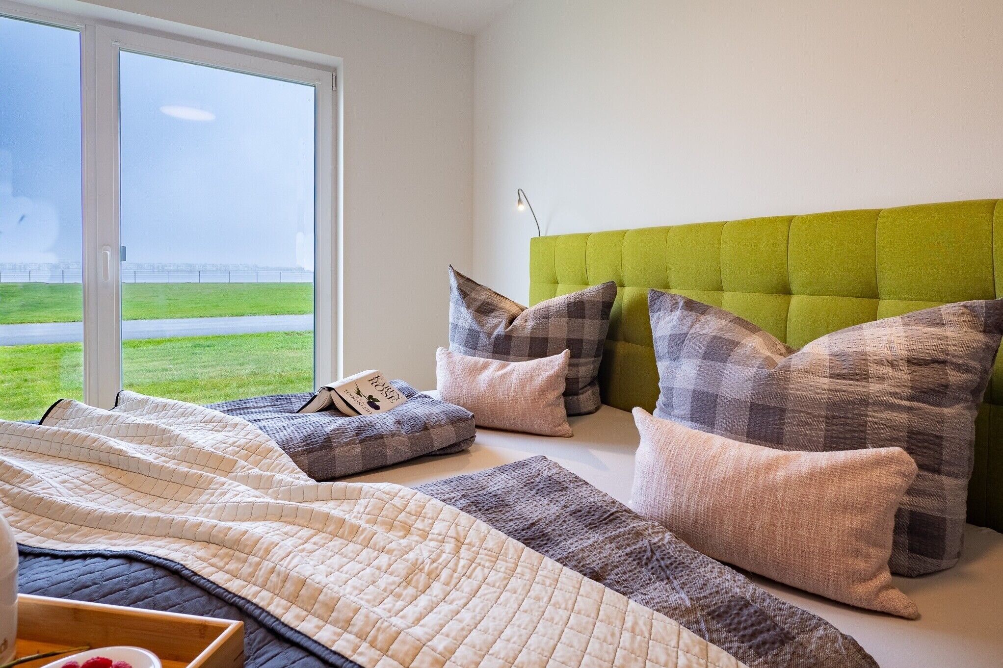 Une chambre à coucher avec une tête de lit recouverte d’un tissu vert, de nombreux coussins, une couvre-lit matelassée et une vue sur un espace verdoyant par une grande fenêtre.