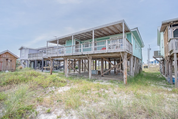 A wooden house with a balcony, situated on stilts over a sandy area.