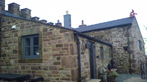 A stone building with a window and a door, a small outdoor seating area, and a potted plant. - 1 Bed Cottage - Sleeps 3 - Garden - Parking - Wifi (Buxton)