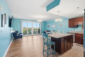 A modern kitchen with a central island, a dining area with chairs, and a large sliding glass door leading to a balcony.