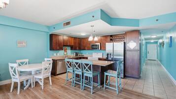 Una cocina con armarios de madera, microondas, estufa y refrigerador. Hay un comedor con mesa blanca y sillas azules.