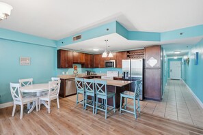 A kitchen with wooden cabinets, a microwave, a stove, and a refrigerator. There is a dining area with a white table and blue chairs.