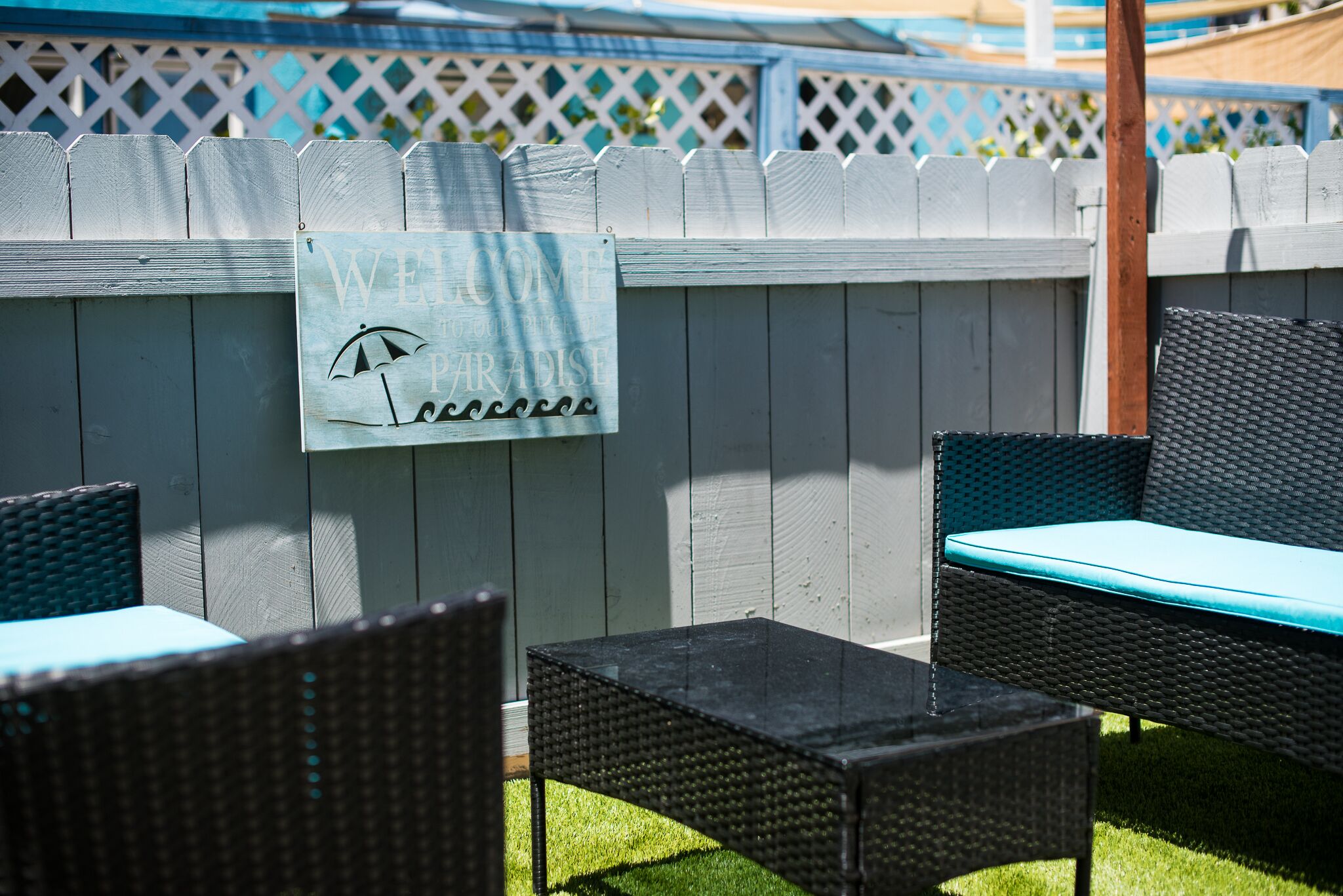 Un espace patio aménagé avec des meubles en osier, une pancarte indiquant «Bienvenue au paradis», et une clôture blanche en bois.