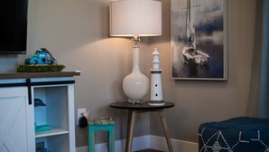 A room with a white lamp on a wooden stand, a small table with a white object, a blue car model, and a painting of a sailboat.