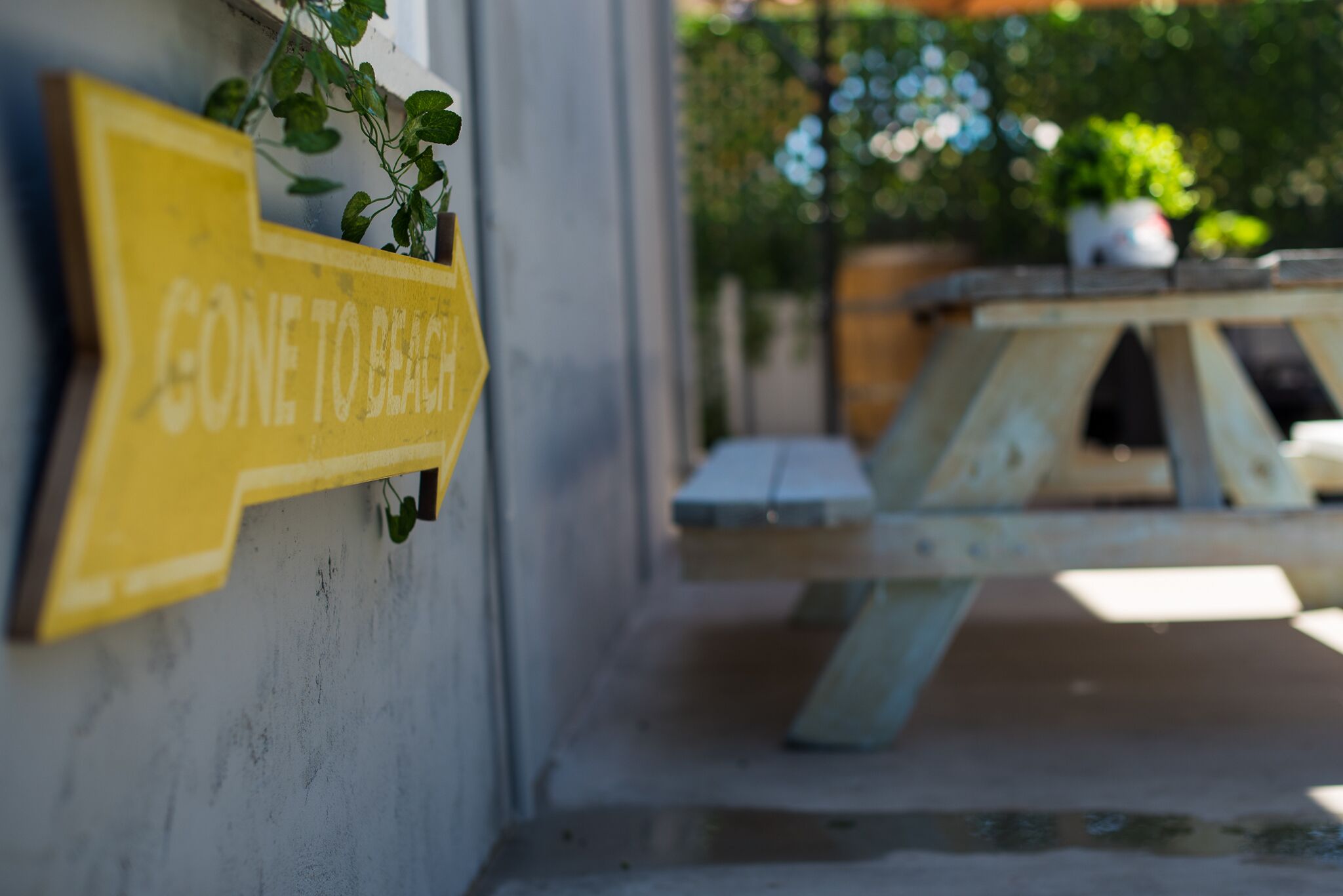 Ein gelbes Hinweisschild mit der Aufschrift "GONE TO BEACH" an einer Betonwand, ein Holztisch und Stühle im Hintergrund sowie Grünpflanzen in der Ferne.