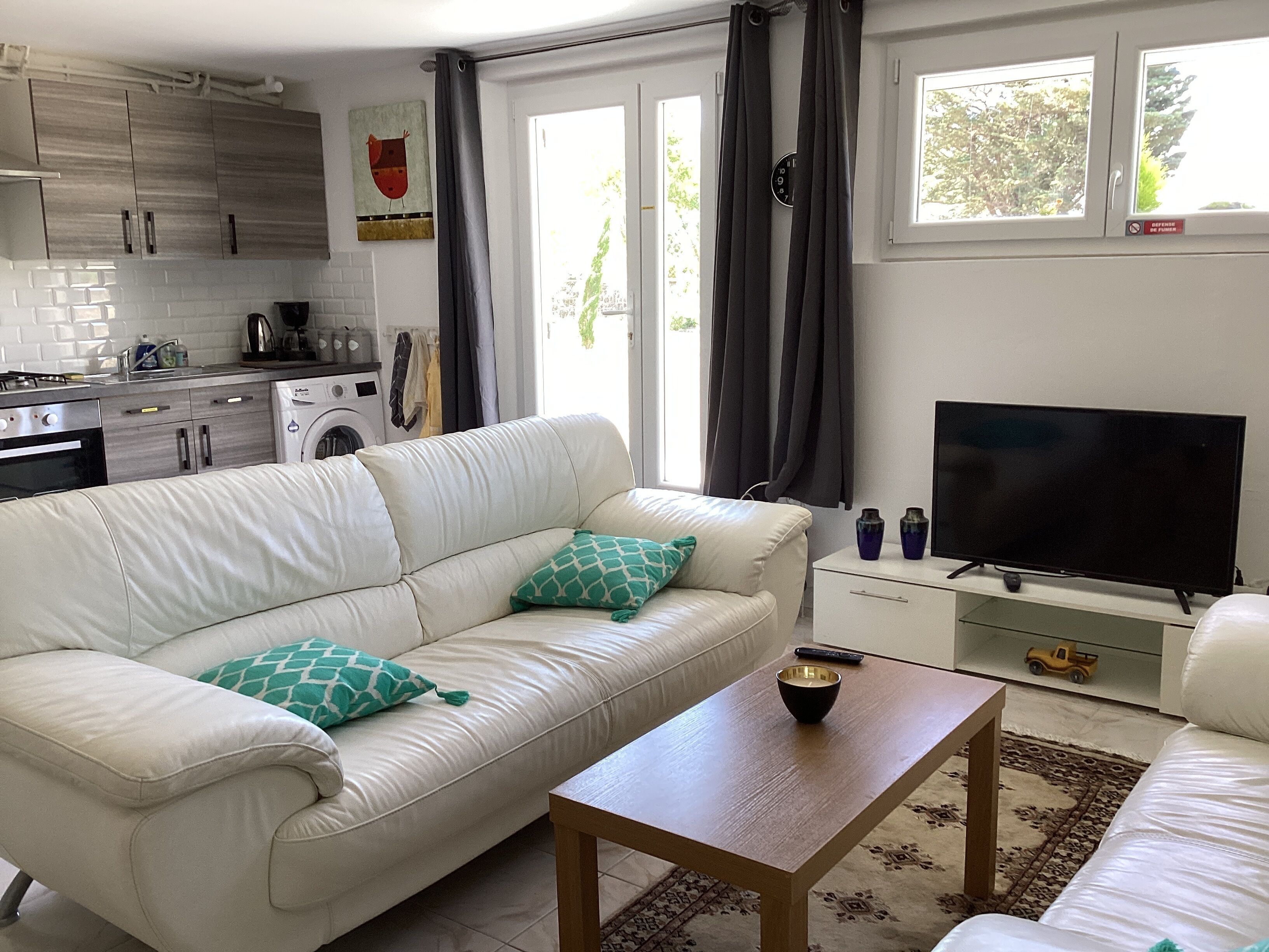 A modern living room with a white leather sofa, a wooden coffee table, a flat-screen TV, and a kitchen area with cabinets and appliances.