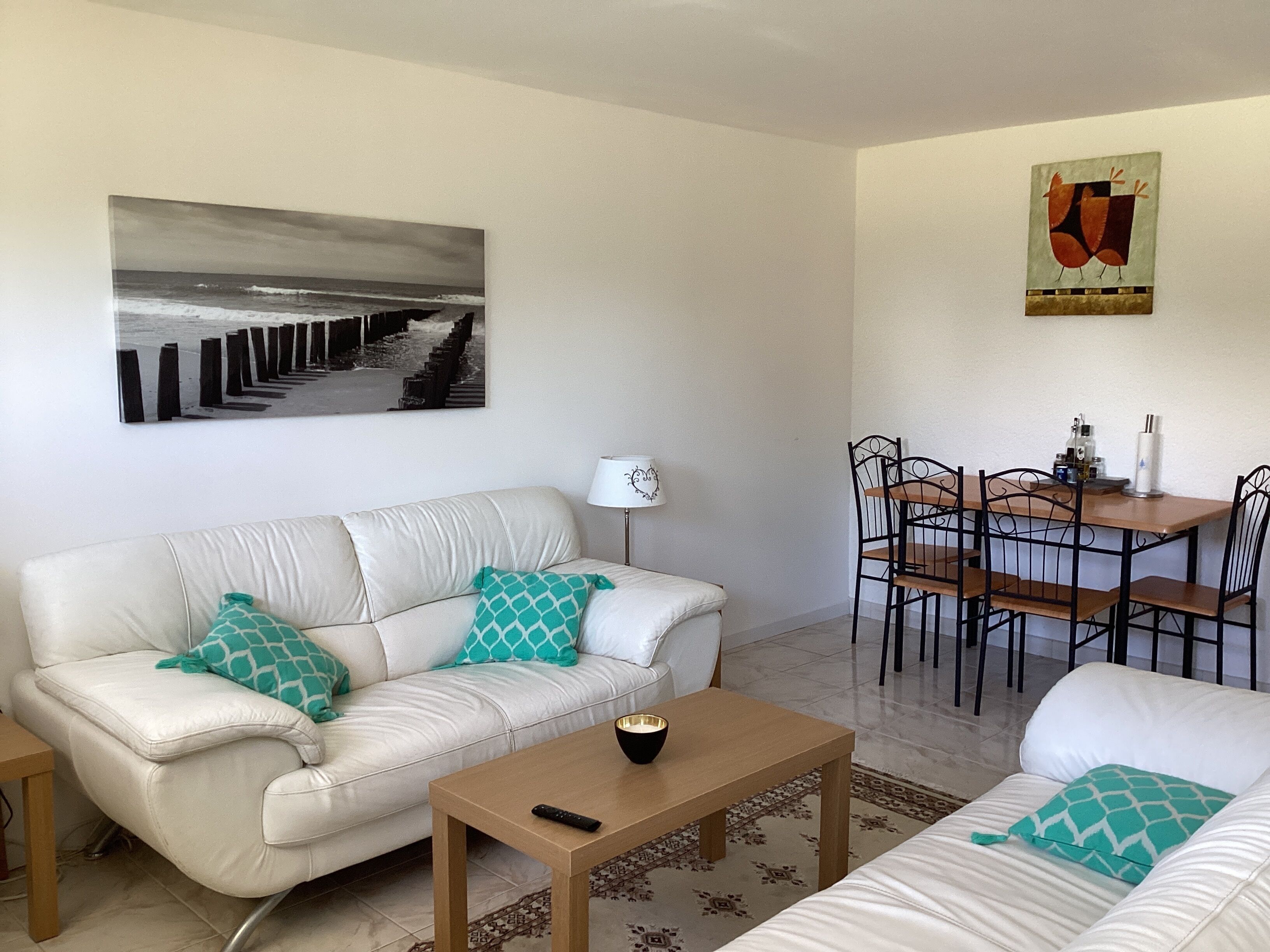 A living room with a white sofa, a coffee table, a dining table with chairs, a lamp, and a painting on the wall.