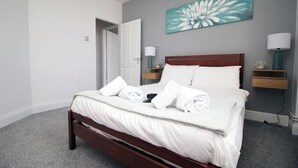 A neatly made bed with white linens and towels, a wooden headboard, a wall-mounted flower painting, bedside lamps, and a small wooden nightstand. - 2 Bed Flat - Sleeps 6 - Parking - Wifi (Swansea)