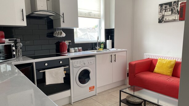 A compact kitchen with a washing machine, a red sofa, and a glass table. - 2 Bed Flat - Sleeps 6 - Parking - Wifi (Swansea)