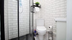 A small, modern bathroom with white subway tile walls, a white toilet, a glass shower enclosure, and a black metal shelf holding a plant and a towel. - 2 Bed Flat - Sleeps 6 - Parking - Wifi (Swansea)