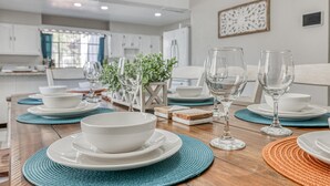 Una mesa de comedor dispuesta con platos, cuencos y copas de vino blancos, rodeada de individuales tejidos y objetos decorativos.