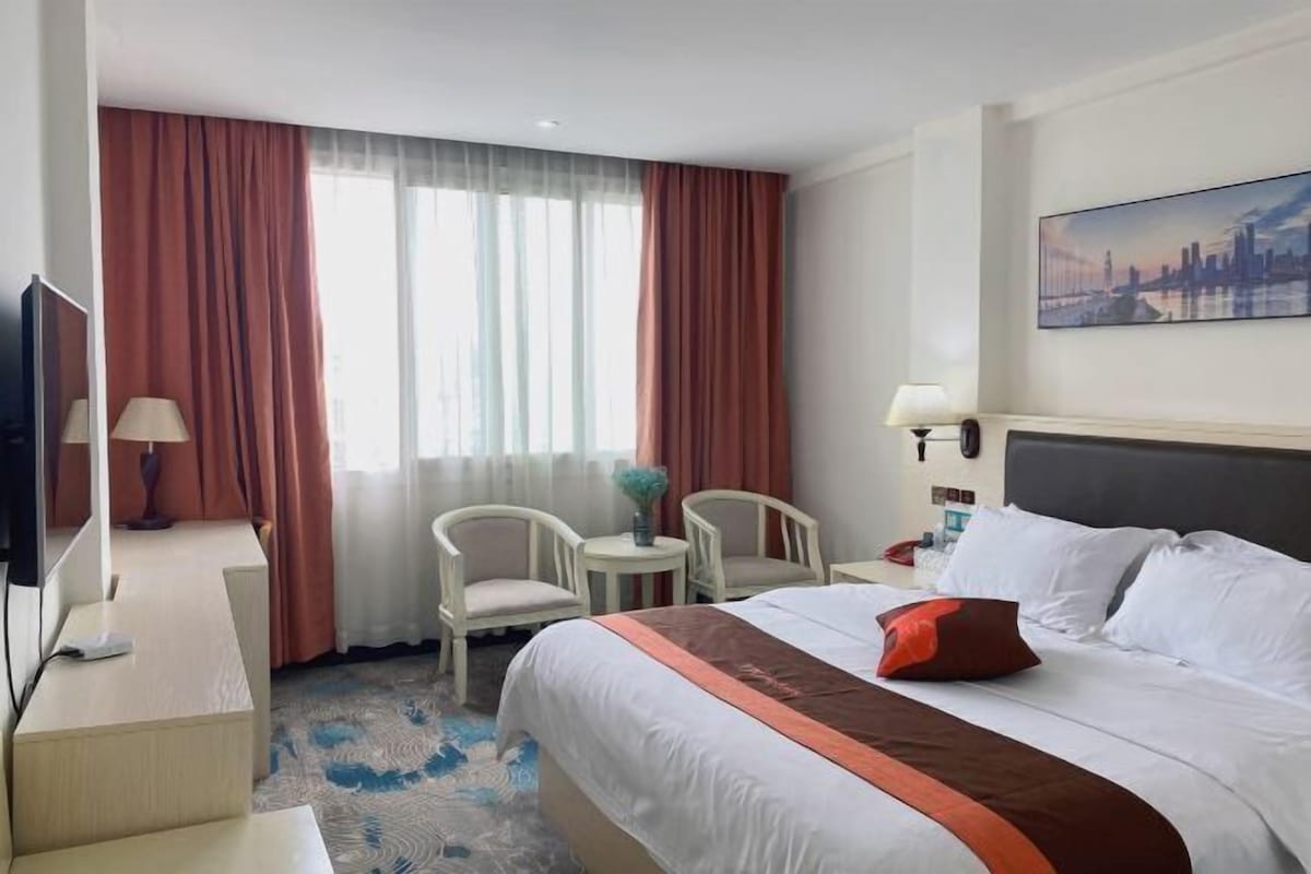 Quarto de hotel com uma cama grande, duas cadeiras, uma mesinha, televisão e um quadro na parede.