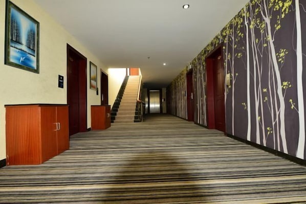 Um corredor de hotel com carpete listrado, portas de madeira e uma escadaria que leva a outro andar.