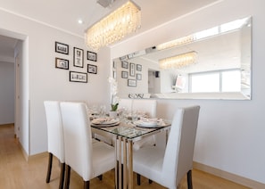 A dining area with a glass table, white chairs, and a chandelier. There are framed pictures on the wall and a large mirror above the table. - Seaview Balcony 4BR/3BA MIRAFLORES (Miraflores)