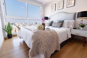 Une chambre avec une grande fenêtre, un lit recouvert d’une couverture moelleuse, une table de chevet avec une lampe, et une vue sur la ville.