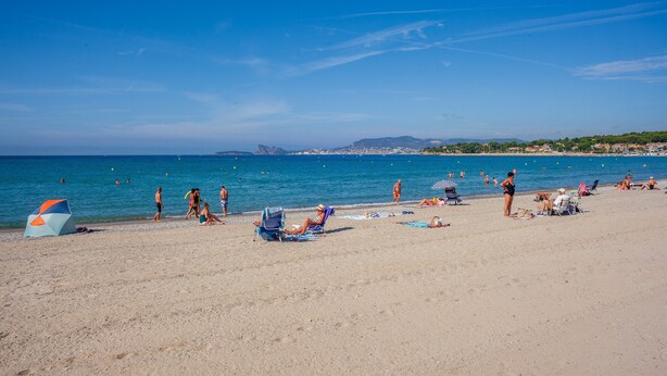 Una playa con gente tomando el sol y nadando, cielo despejado y montañas a lo lejos.