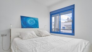 Ein Schlafzimmer mit einem Bett, einem Fenster mit blauem Rahmen, einer weißen Wand und einem Gemälde.