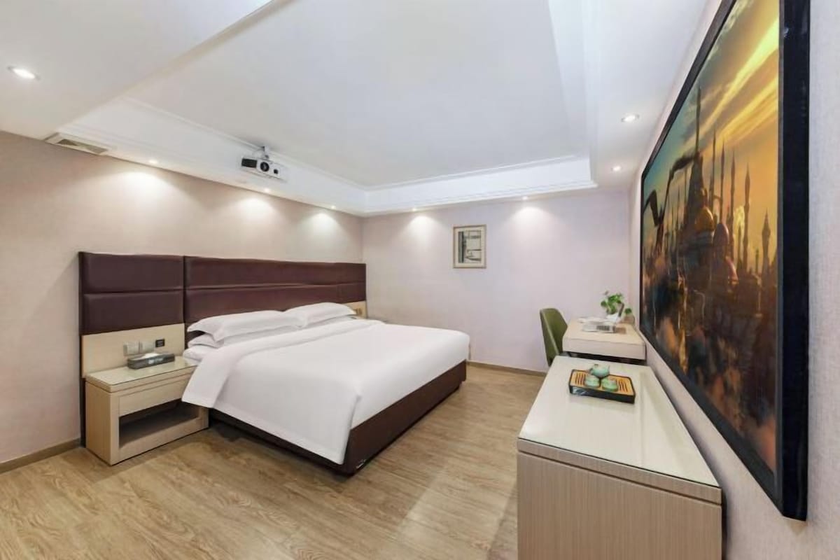 A hotel room with a bed, bedside table, desk, chair, and a painting on the wall.