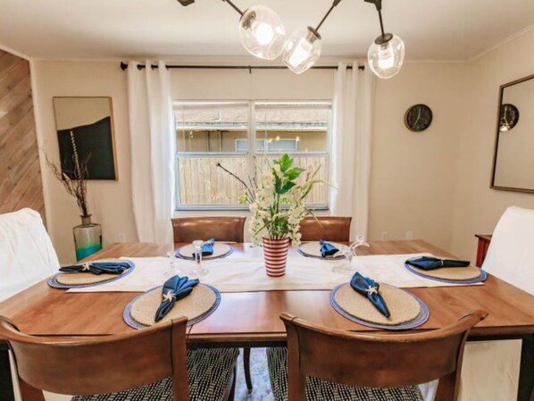 A dining area with a wooden table set for four, surrounded by chairs. There is a potted plant in the center, and the room has a window with curtains. - HAPPY FUN BLUE HOUSE for 8 people kids, pets friendly, 3 miles from the beach🏖️ (Venice)