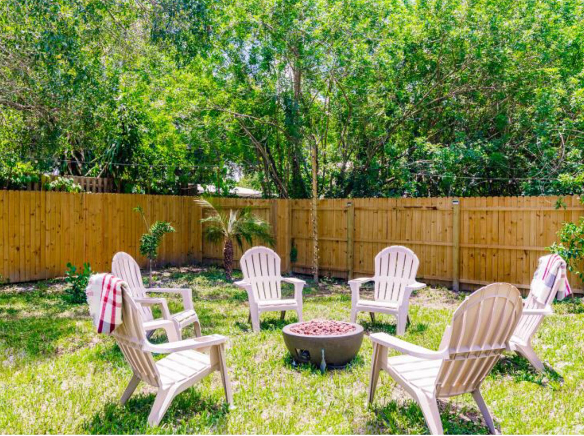 Un jardin avec une clôture en bois, de l’herbe verte et des chaises en plastique blanches disposées autour d’un foyer.