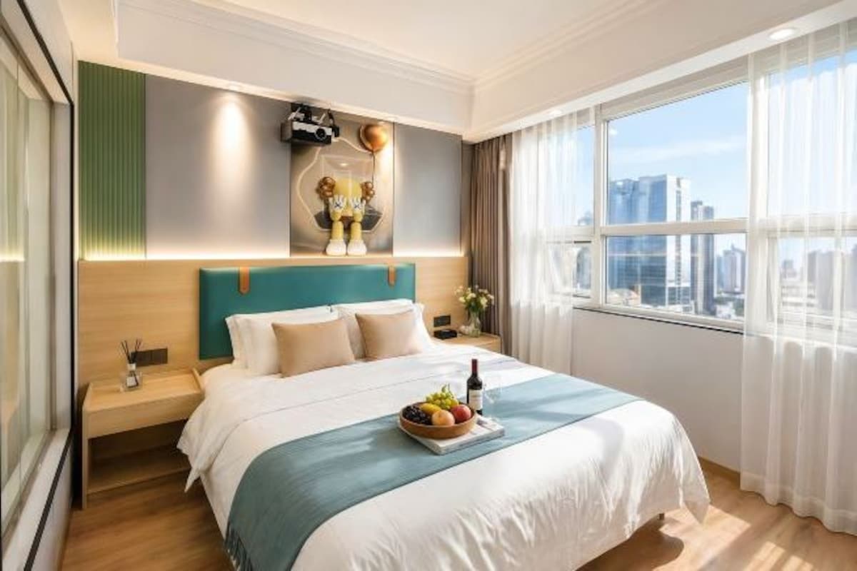Una habitación de hotel con cama, mesita de noche, ventana con vista a la ciudad y una lámpara fijada en la pared.