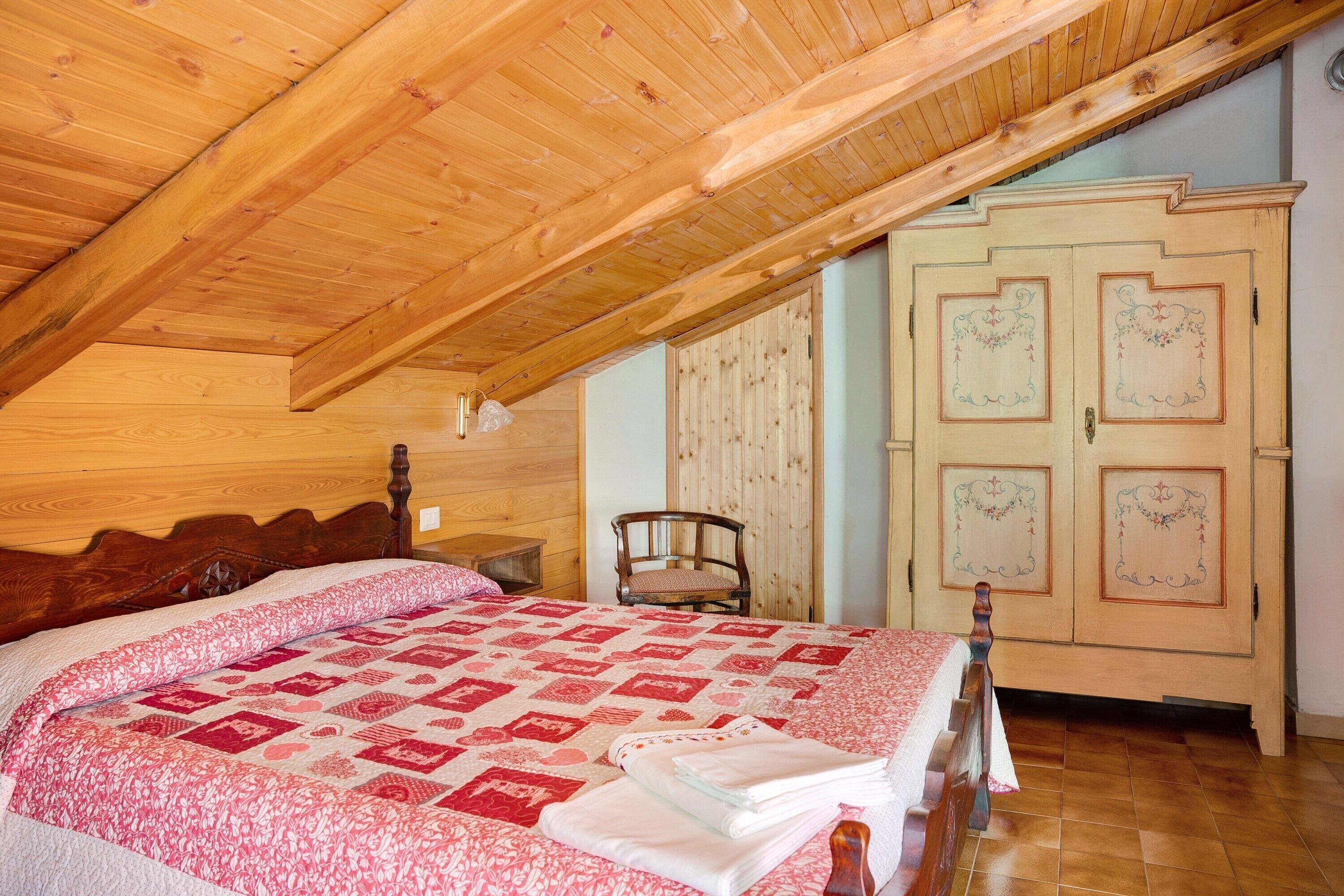 A bedroom with a wooden ceiling, a bed with a patterned quilt, a wooden chair, and a wardrobe with decorative panels.