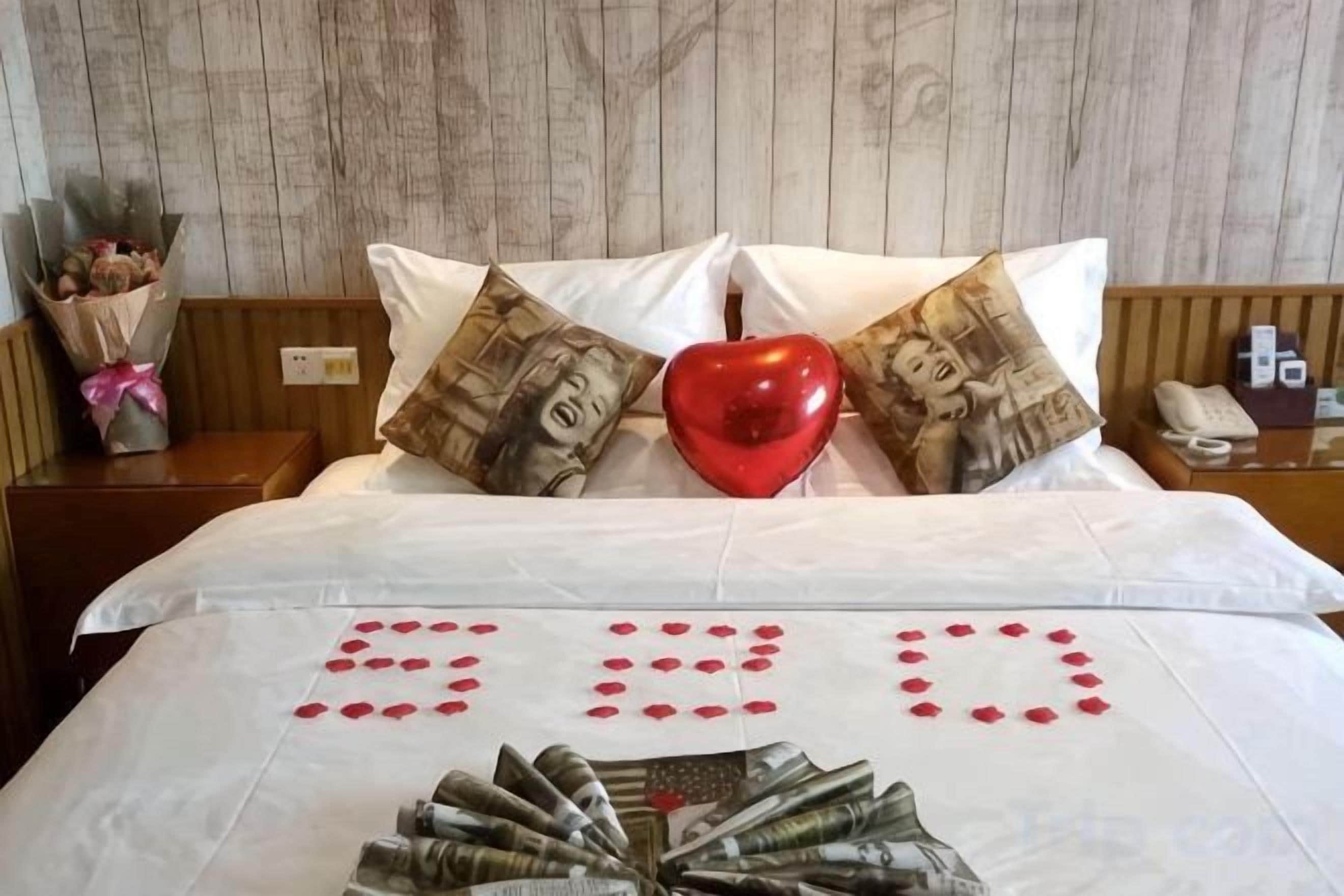 A bed with a heart-shaped pillow, a bouquet of flowers, and a red heart on the bedspread.