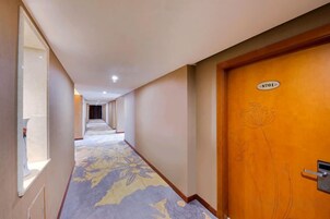 A hotel corridor with a carpeted floor, a wooden door with a number, and a window on the left.