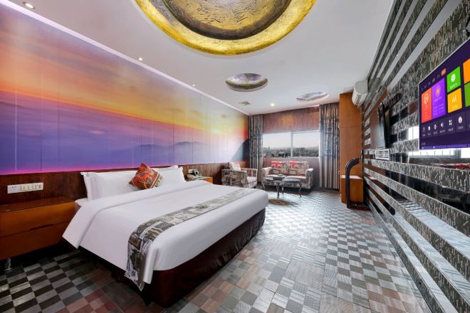A hotel room with a large bed, a checkered floor, a wall with a sunset mural, a TV, and a balcony with a view.