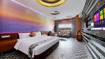 A hotel room with a large bed, a checkered floor, a wall with a sunset mural, a TV, and a balcony with a view.